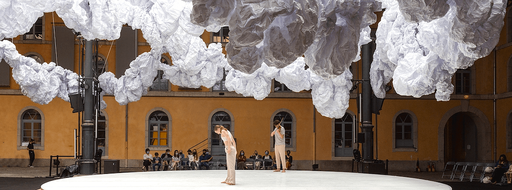 A contemporary dance performance taking place on a circular stage, surrounded by large, cloud-like fabric formations, with an audience in the background watching.